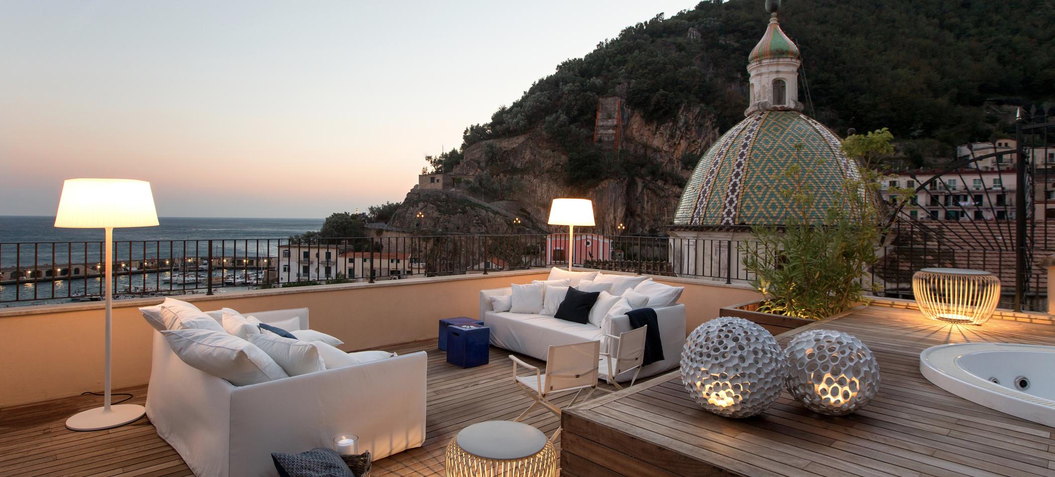 Panoramisk takterrasse i Cetara med hvite utem&oslash;bler, tregulv, stemningsfull belysning og utsikt mot havet og den keramikkkledde kirkekuppelen ved solnedgang.