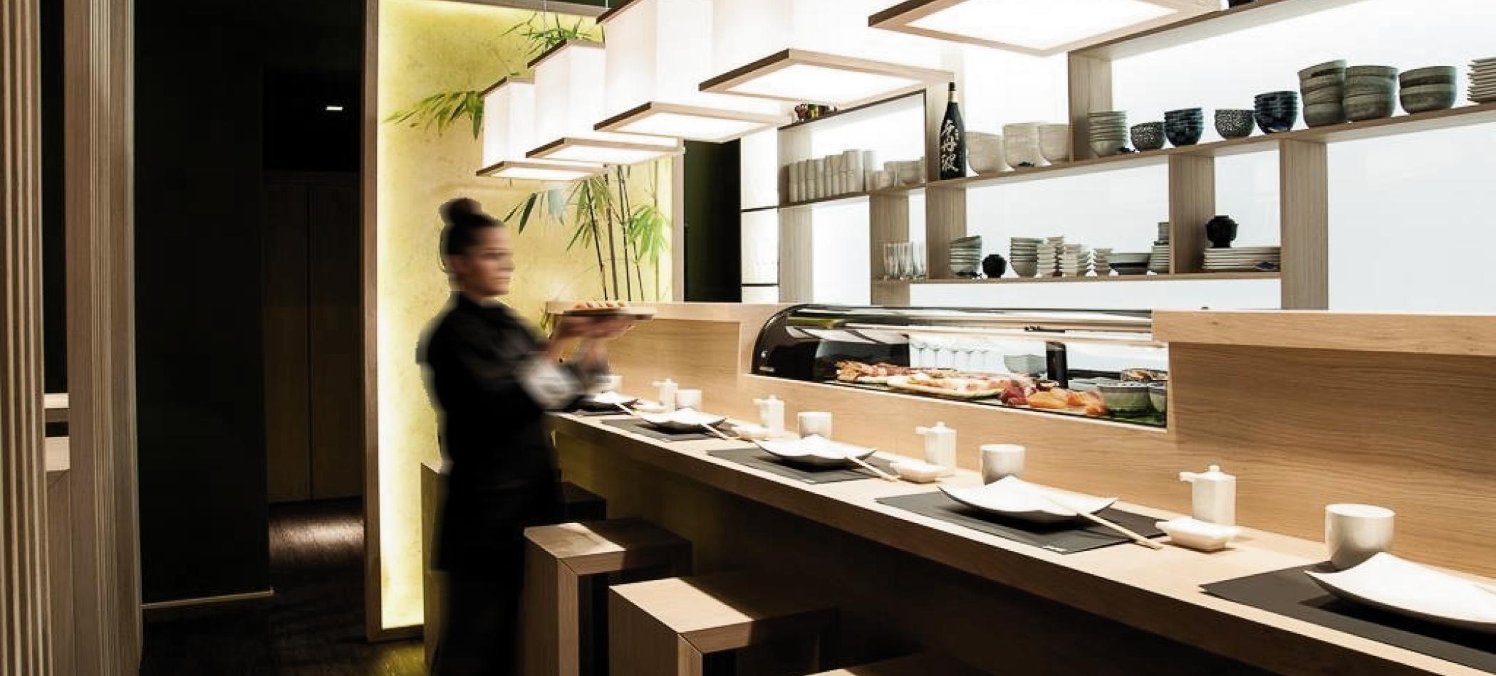 Sushi-bar med lang trebenk, sitteplasser ved disken og gjester som f&oslash;lger sushi-tilberedningen i et intimt rom.