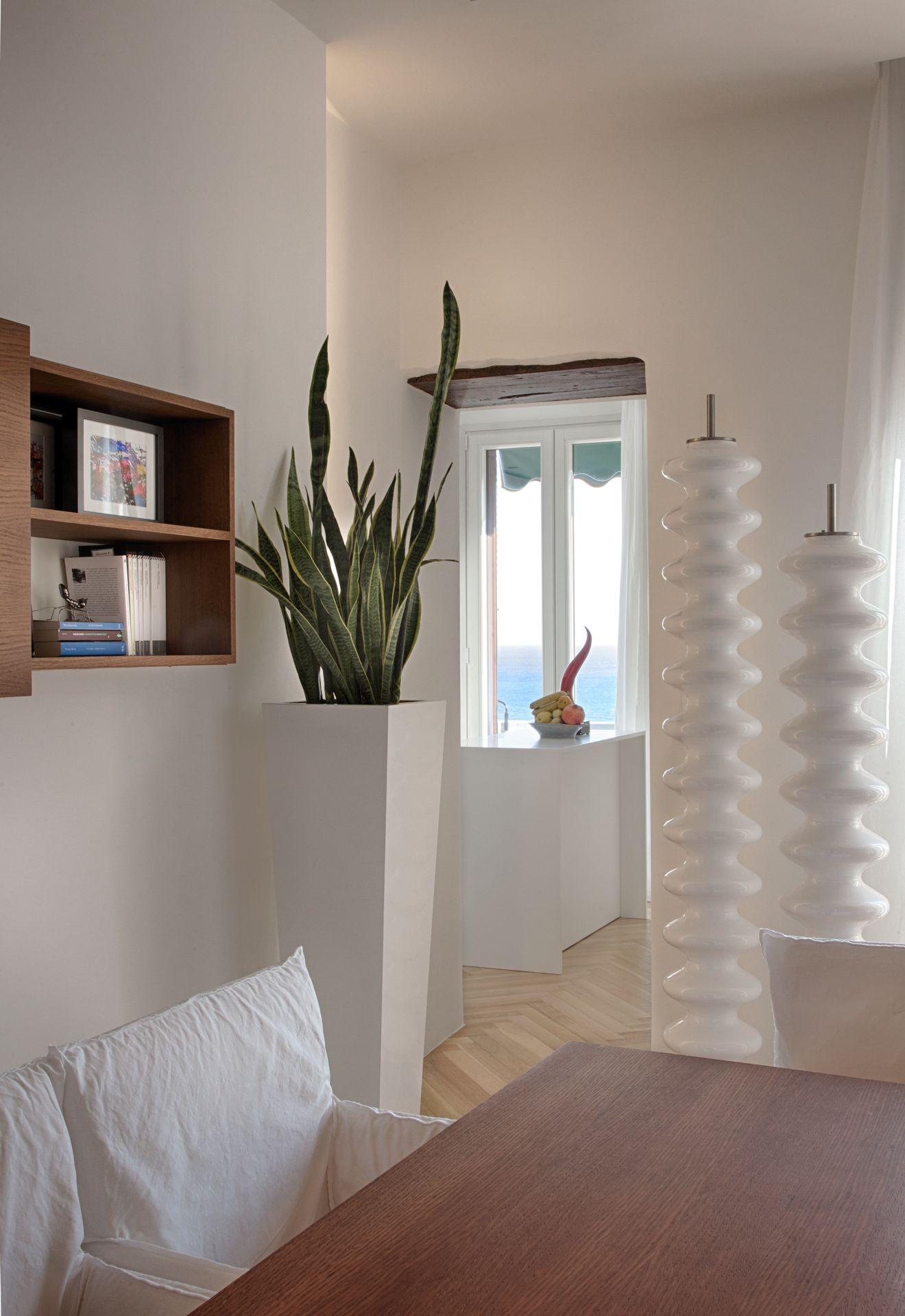 Prosjekt for interi&oslash;r og eksteri&oslash;r av Ernesto Fusco p&aring; Amalfikysten. Overgang mellom kj&oslash;kken og oppholdsrom med hvite, rene flater, spesiallagde sokler i hvit finish, innebygde plantekasser, fiskebeinsparkett i lys eik og utsikt mot havet gjennom vinduet.