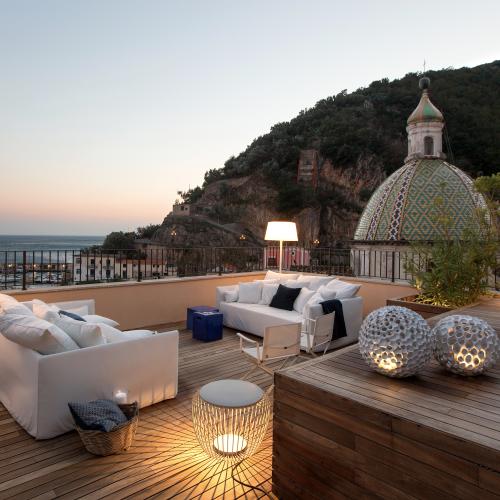 Panoramisk takterrasse i Cetara med hvite utem&oslash;bler, tregulv, stemningsfull belysning og utsikt mot havet og den keramikkkledde kirkekuppelen ved solnedgang.